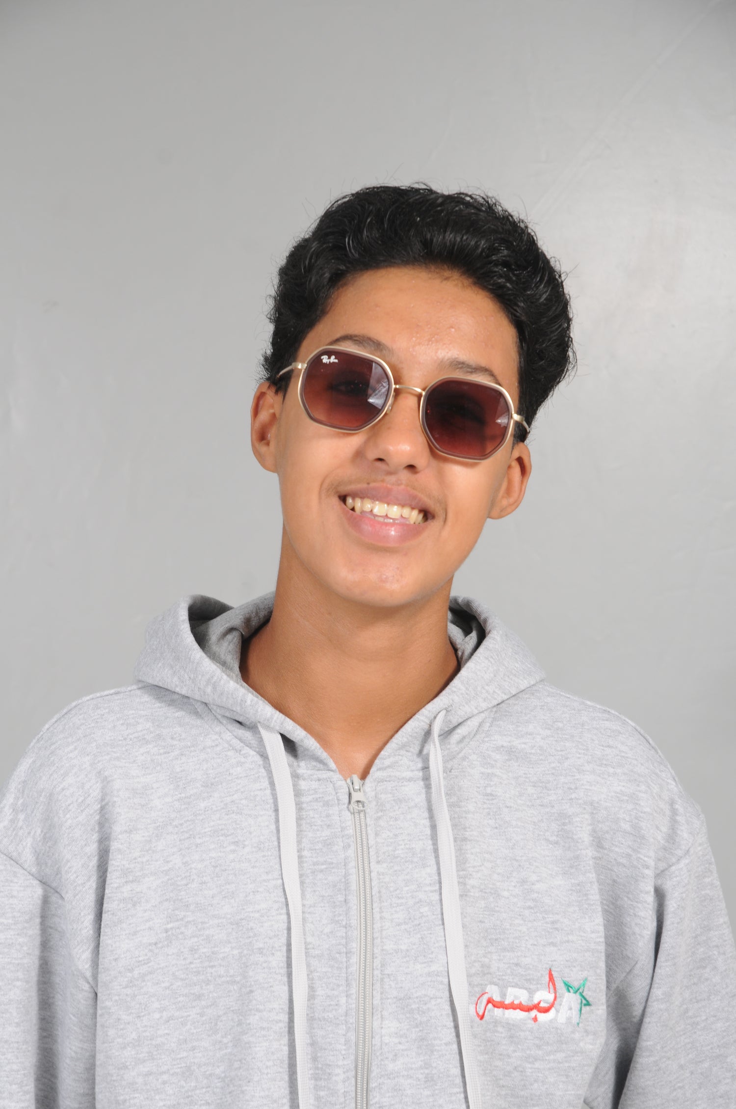 Person wearing sunglasses and a gray hoodie with a logo on a plain background